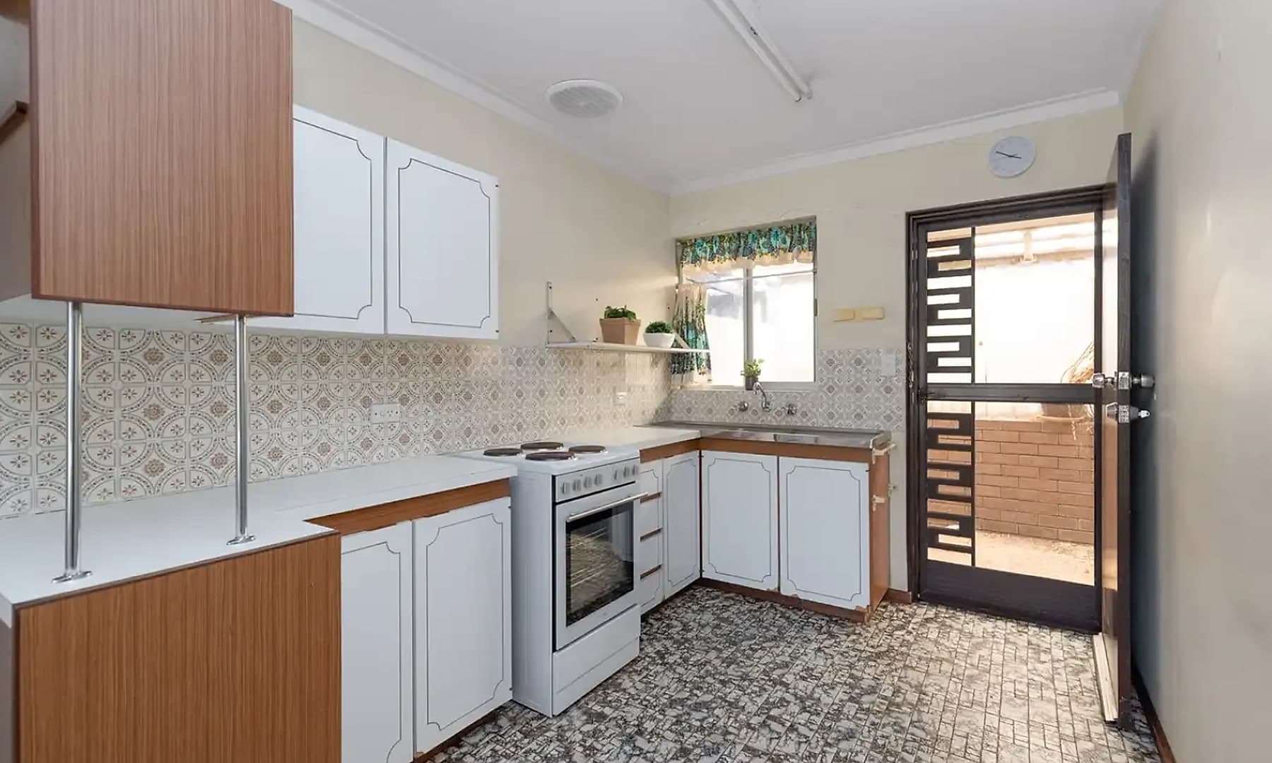 The Maylands kitchen before the renovation, the original early 1980s fitout with white and timber laminate cabinets, a patterned tiled splashback, a freestanding electric range and worn vinyl flooring.