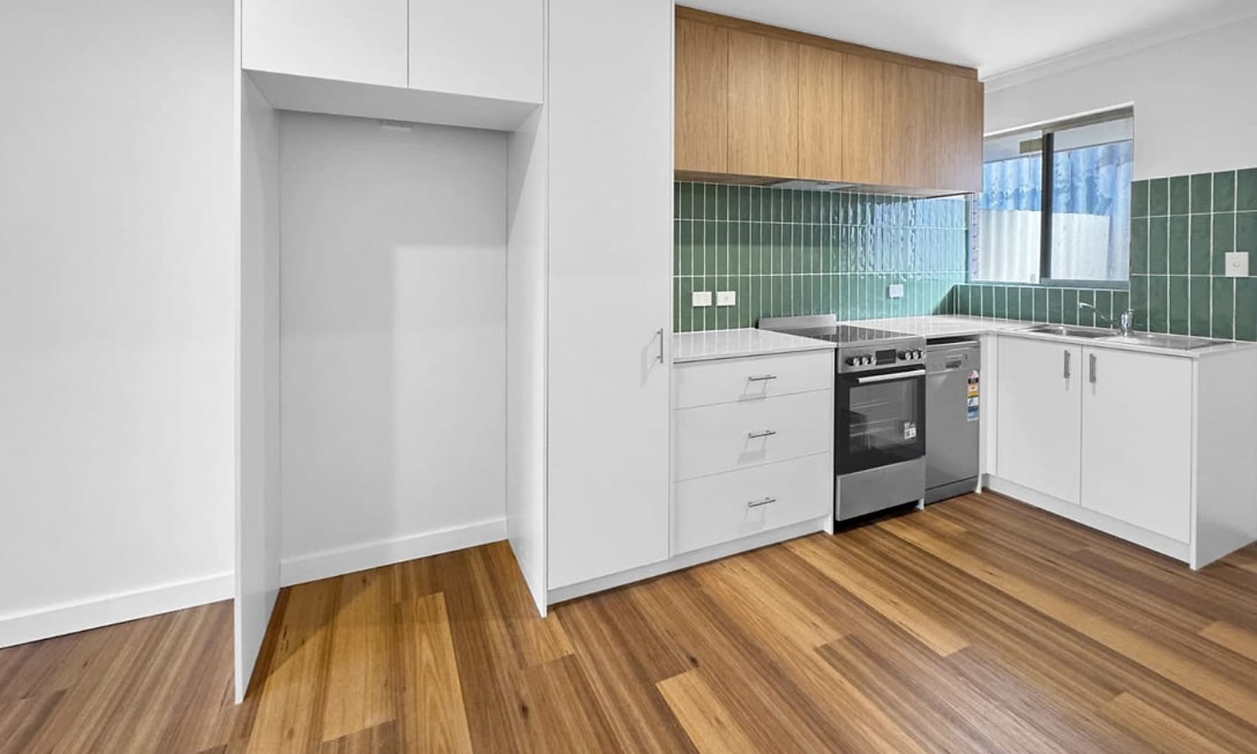 The same kitchen after the renovation, rebuilt with white cabinetry and oak overheads, a green tiled splashback, timber floors, a new oven and a dishwasher.