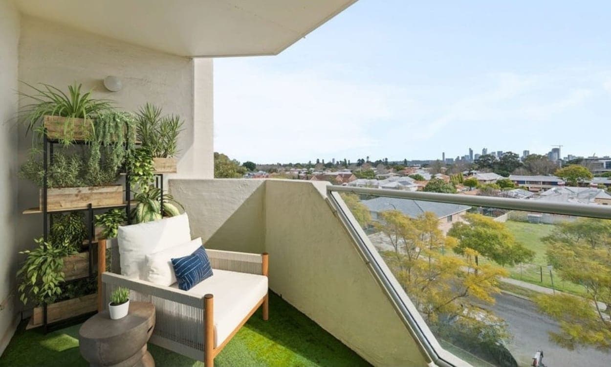 The balcony of the Victoria Park apartment, looking out over rooftops and a park to the city skyline, with a two seater lounge and a shelf of plants.