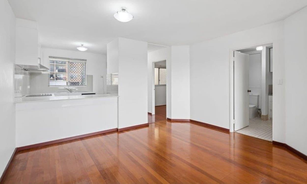The open plan living area, with polished timber floors, white walls, a kitchen bench along one wall and the bathroom off to the side.
