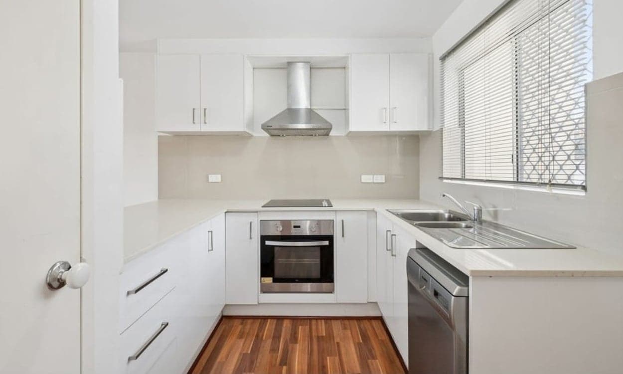 The kitchen, renovated in white with stone tops, an electric cooktop under a stainless canopy, a wall oven and a dishwasher.