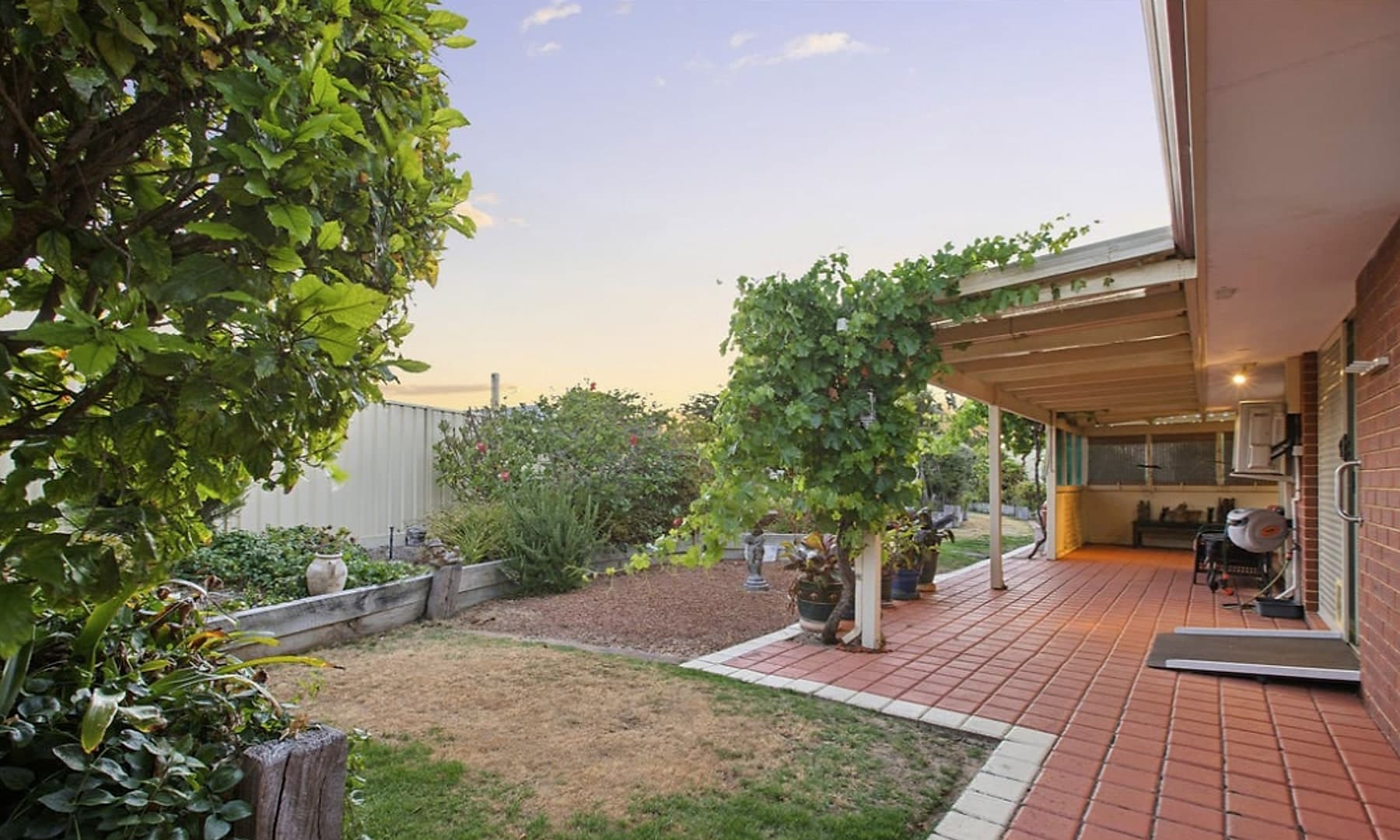 The back garden at dusk, with a paved patio under a pergola, raised beds and a lawn.